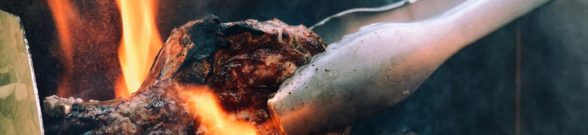 a grill with flames and tongs turning some charred meat.
